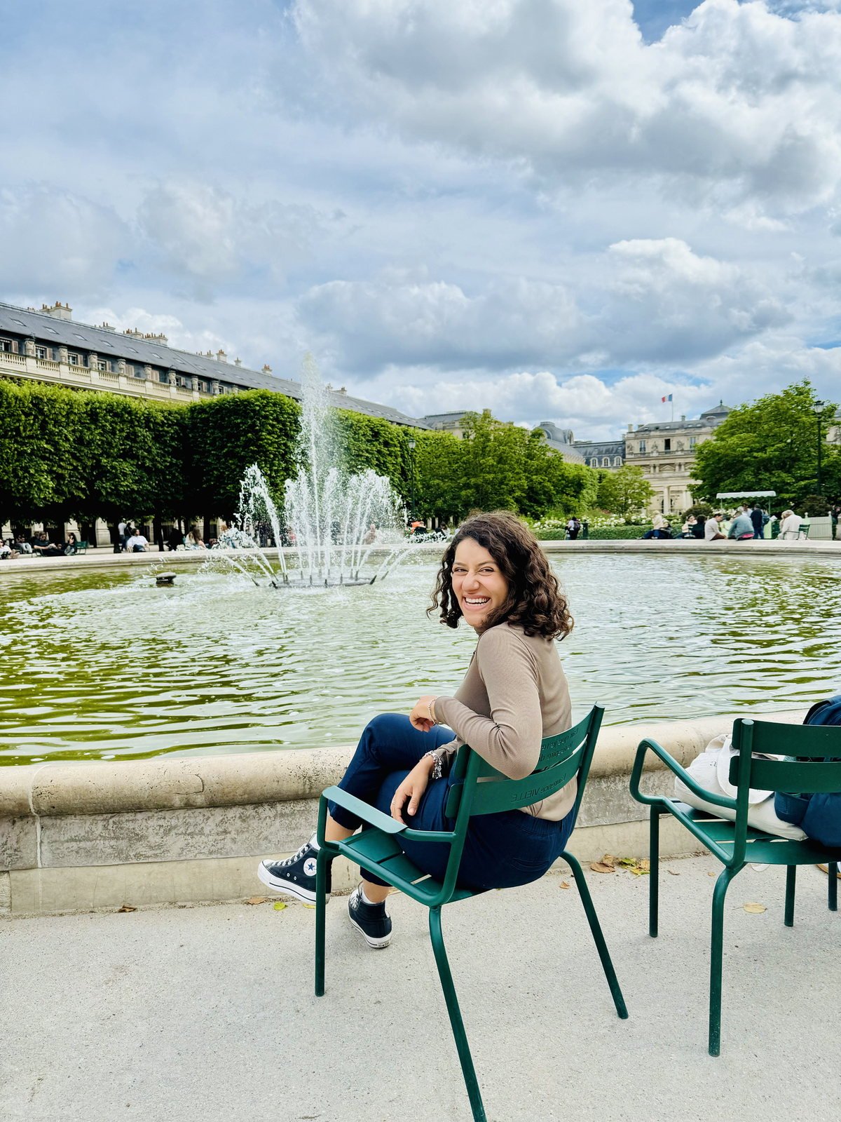 Mari sentada numa cadeira de jardim em Paris, com uma fonte ao fundo